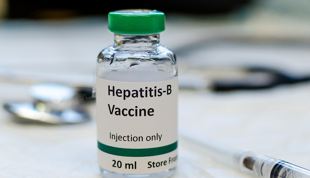 A 20ml vile of the hepatitis B vaccine with a needle next to it. A stethoscope is blurred in the background.