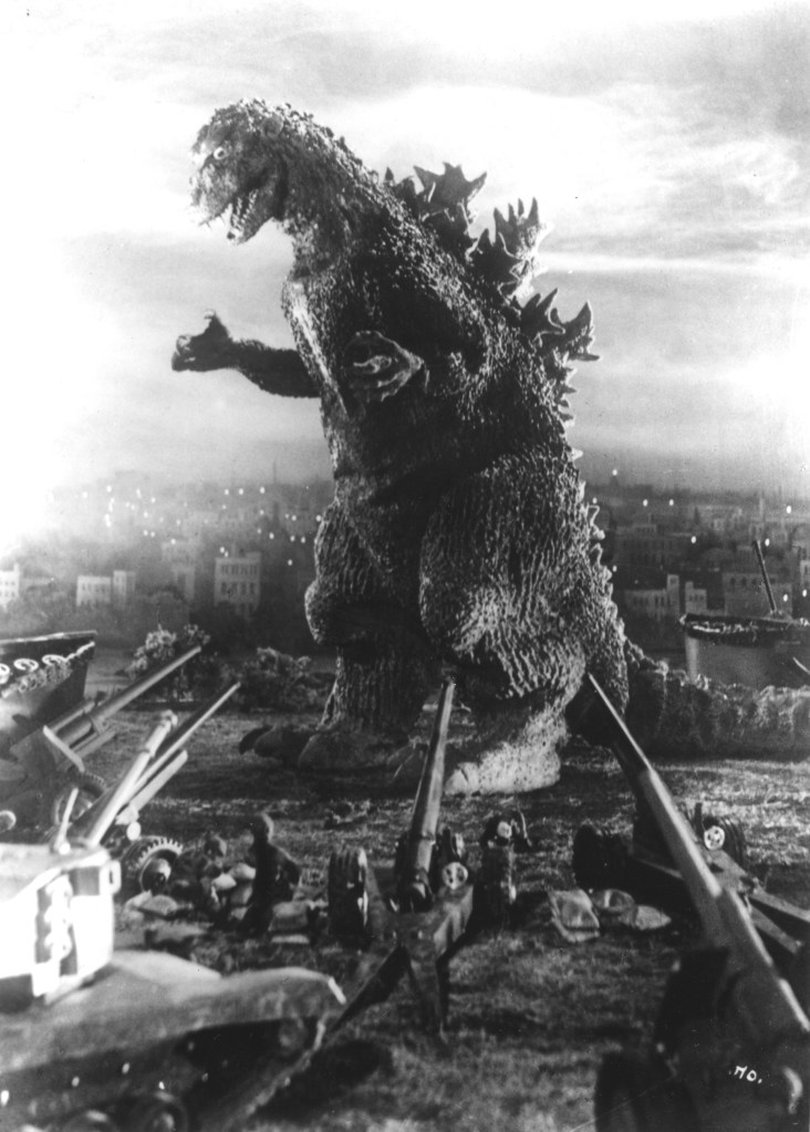 A screen capture from the black and white Japanese movie Godzilla. The monster is surrounded by army men and a larger number of guns and tanks.