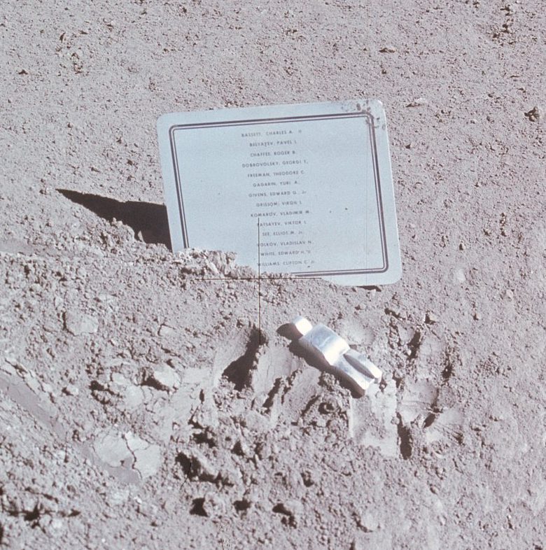 An image of the fallen astronaut on the grey chalky surface of the moon. The fallen astronaut is a aluminium figure. Behind it is a plaque of all the names of those who have died in space.