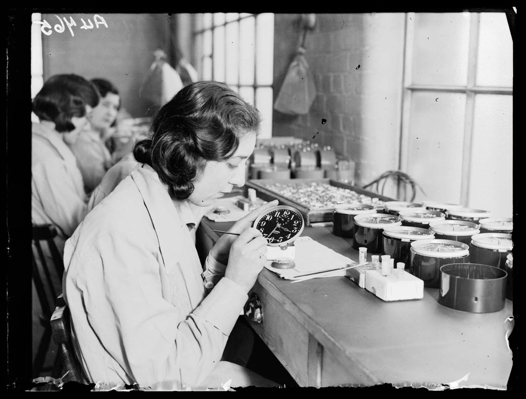 Black and white photo of a woman using glow in the dark radioactive radium-based paint to paint watch dials. She is surrounded by unfinished clocks. Two women are doing the same in the background.